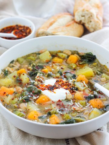 Close shot of a bowl of butternut white bean soup with Swiss chard and a small bowl of crispy chili onion oil and crusty bread of in the background.