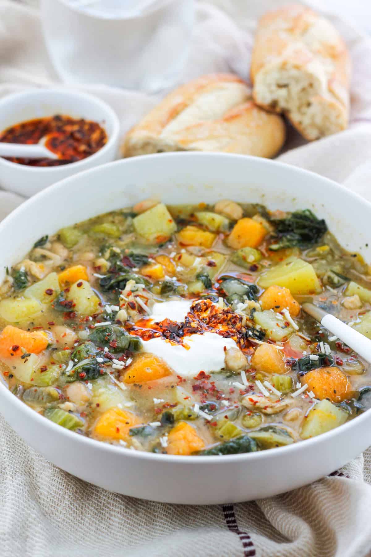 Close shot of a bowl of butternut white bean soup with Swiss chard and a small bowl of crispy chili onion oil and crusty bread of in the background.