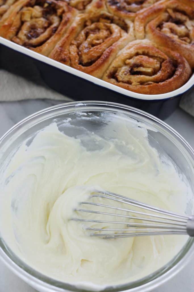 Overhead shot of cream cheese topping for caramel apple cinnamon rolls in a glass bowl with the baked rolls in the background.