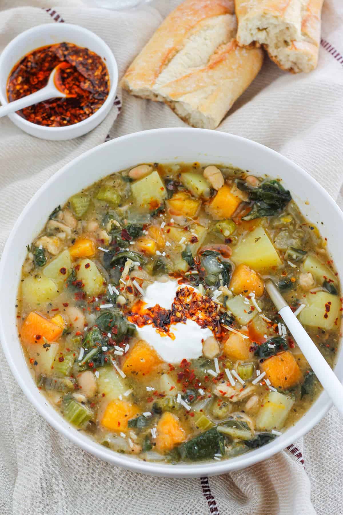 Overhead shot of a bowl of butternut white bean soup with Swiss chard and a small bowl of crispy chili onion oil and crusty bread of in the background.