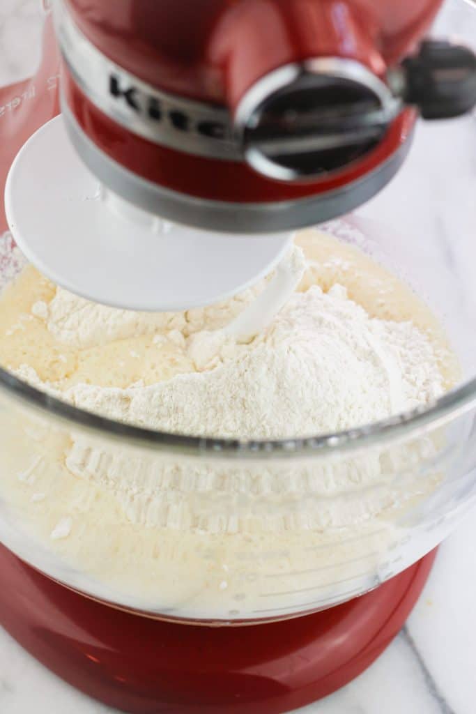 Overhead shot of the flour mixture being added to the liquid mixture of the cinnamon roll dough in a Kitchenaid stand mixer.