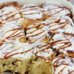 Shot of caramel apple cinnamon roll in a baking dish topped with cream cheese glaze and drizzled wth caramel sauce.