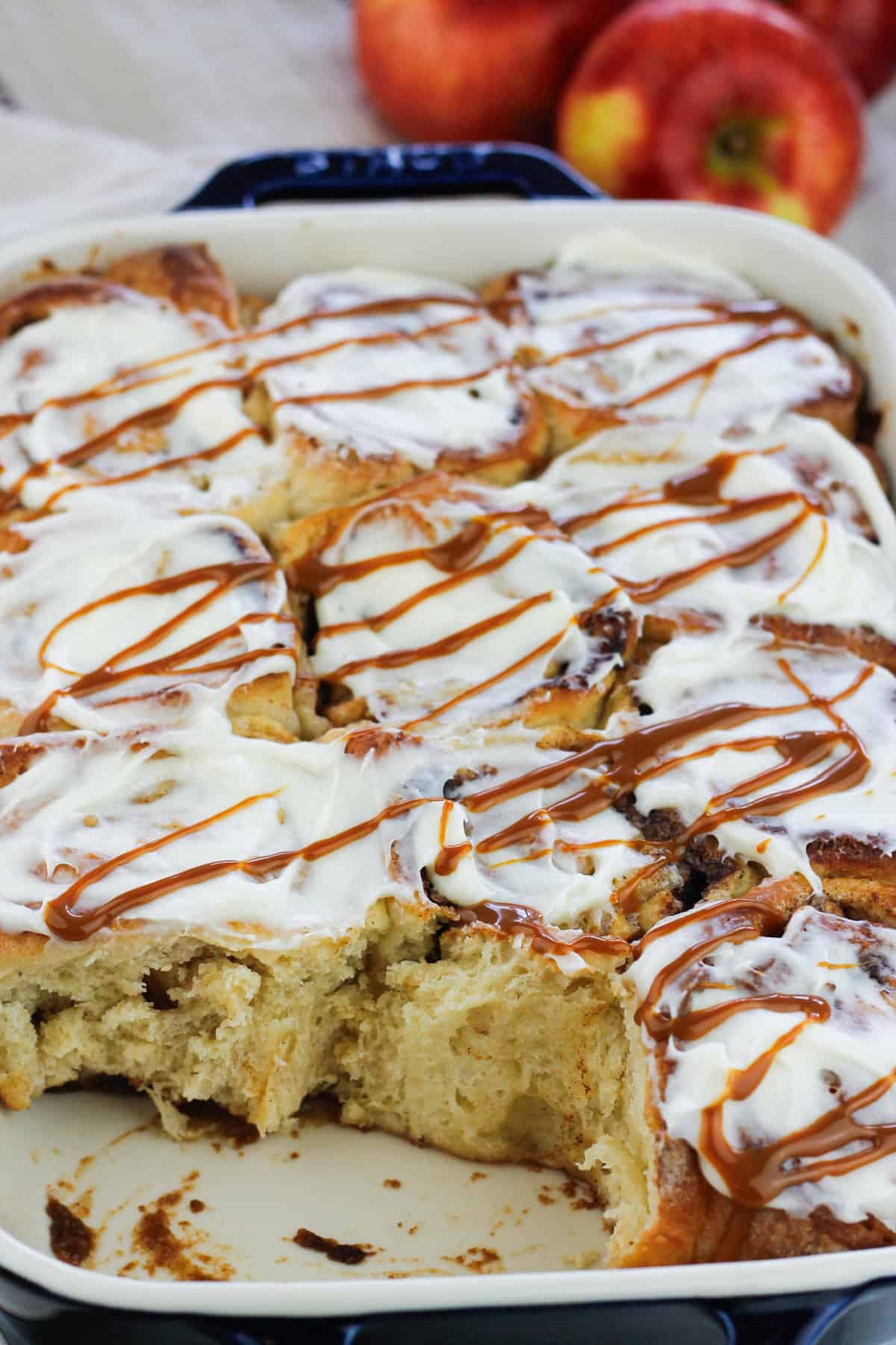 Shot of caramel apple cinnamon roll in a baking dish topped with cream cheese glaze and drizzled wth caramel sauce.