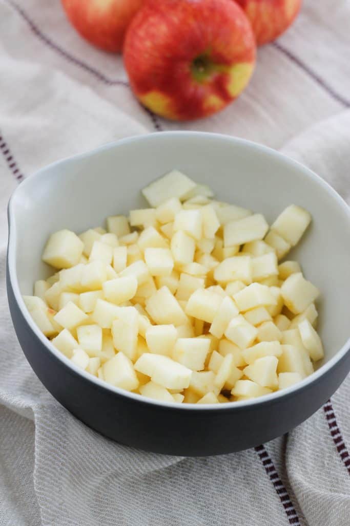 Shot of peeled, diced apples in a bowl with three red apples in the background.