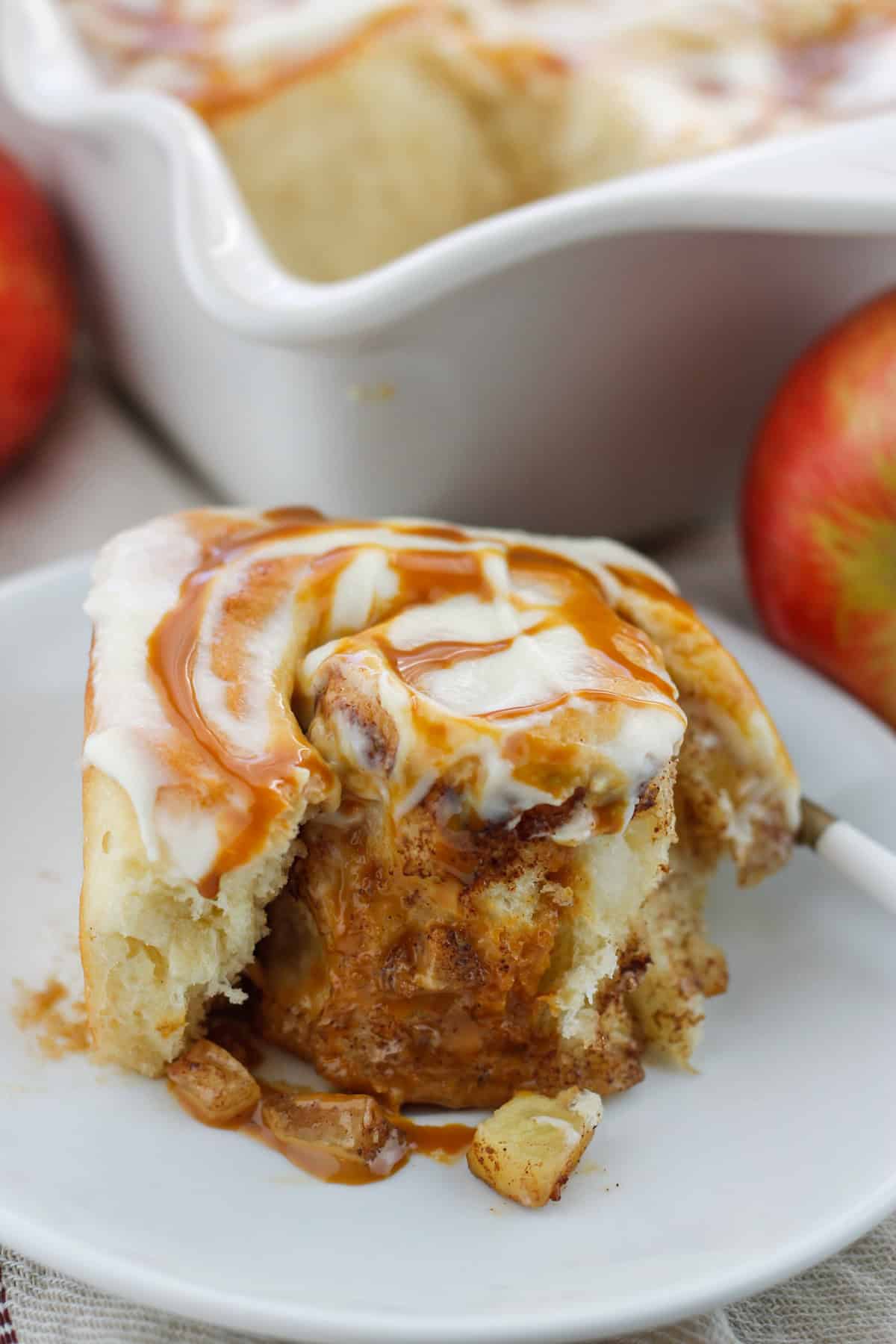 Very close shot of one caramel apple cinnamon roll on a small plate partially unrolled with baked apple bits and caramel on plate with baking dish and in the background.