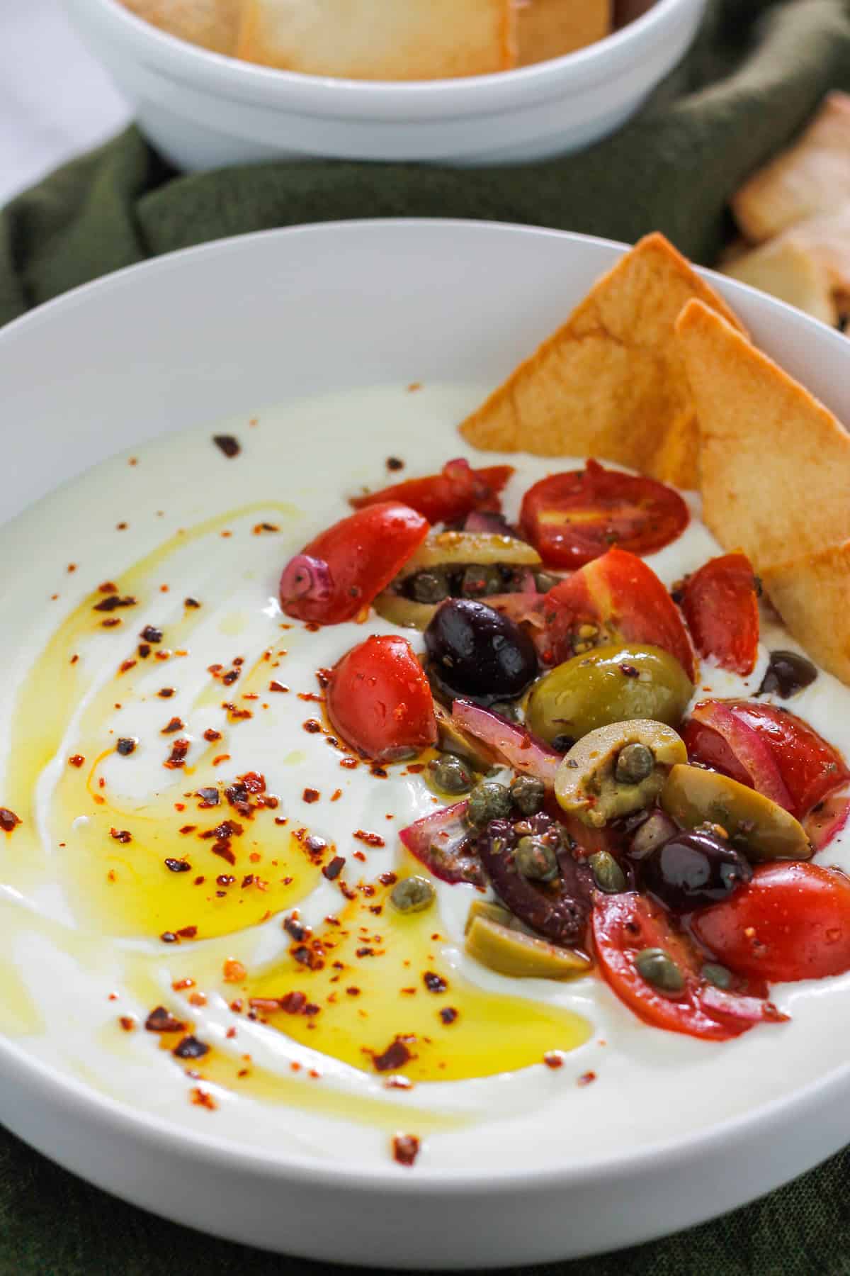 A bowl filled with whipped feta garnished with olive oil, aleppo pepper flakes, tomato olive salad, and crisp pita chips.