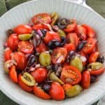 Angle overhead shot of tomato and olive salad in a bowl placed on top of a dark green kitchen towel.