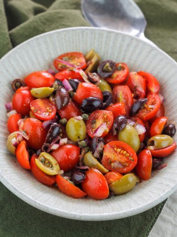 Angle overhead shot of tomato and olive salad in a bowl placed on top of a dark green kitchen towel.