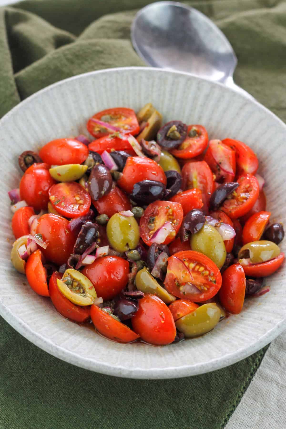 Angle overhead shot of tomato and olive salad in a bowl placed on top of a dark green kitchen towel.