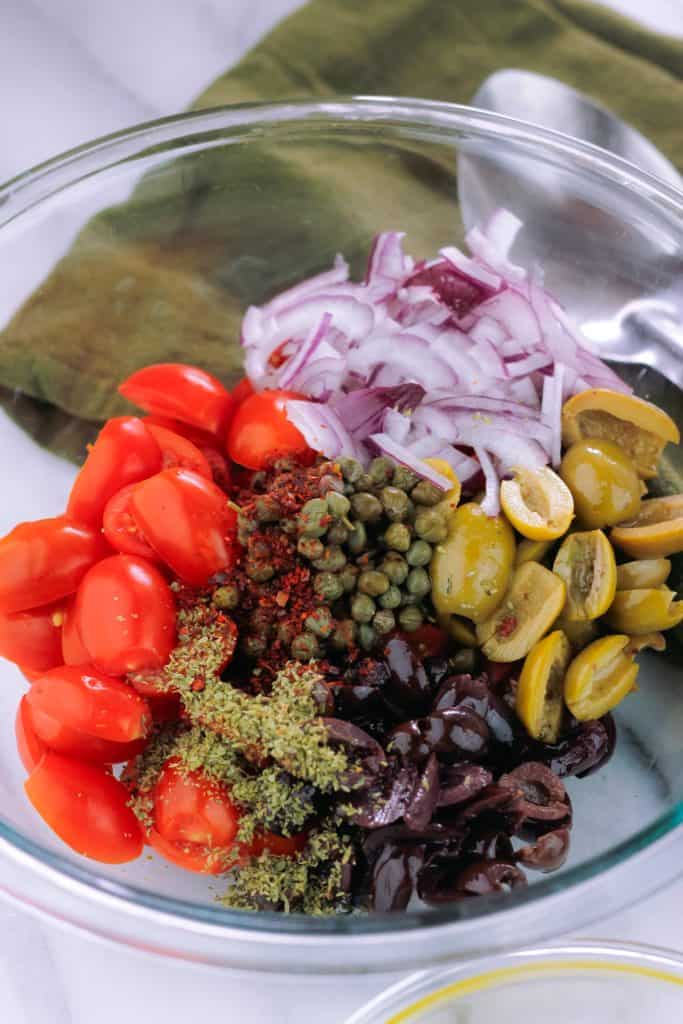 Bowl filled with tomato and olive salad ingredients next to a dark green kitchen towel.