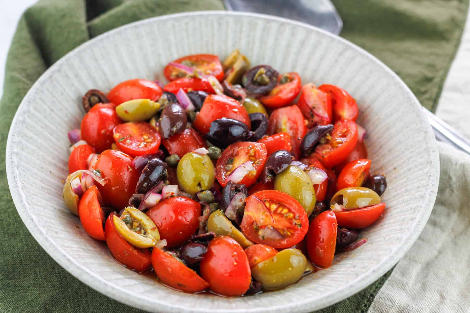 Close-up shot of tomato and olive salad in a bowl placed on a dark green kitchen towel.