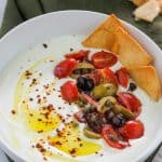 Overhead angled shot of a bowl filled with whipped feta garnished with olive oil, aleppo pepper flakes and some tomato and olive salad with crisp pita chips at the top right side of the bowl.