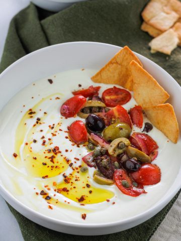 Overhead angled shot of a bowl filled with whipped feta garnished with olive oil, aleppo pepper flakes and some tomato and olive salad with crisp pita chips at the top right side of the bowl.