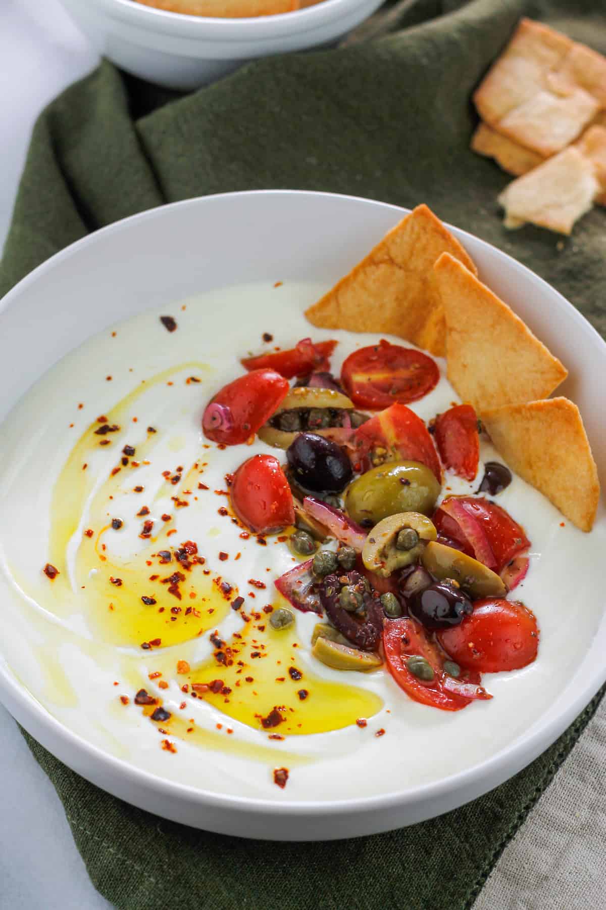 Overhead angled shot of a bowl filled with whipped feta garnished with olive oil, aleppo pepper flakes and some tomato and olive salad with crisp pita chips at the top right side of the bowl.