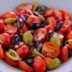 Very close shot of a bowl filled with tomato and olive salad sitting on a dark green kitchen towel with a spoon at the top right side of the bowl.