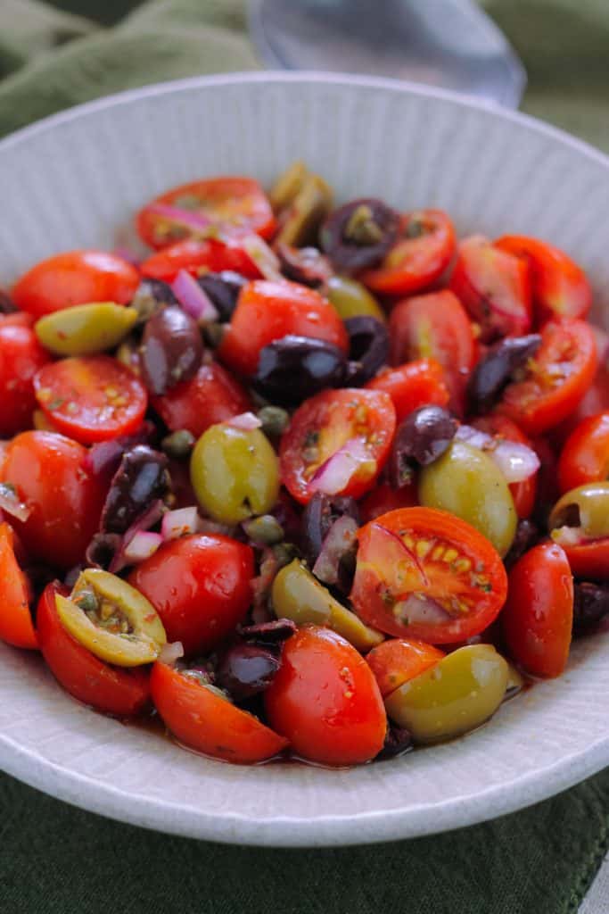 Very close shot of a bowl filled with tomato and olive salad sitting on a dark green kitchen towel with a spoon at the top right side of the bowl.