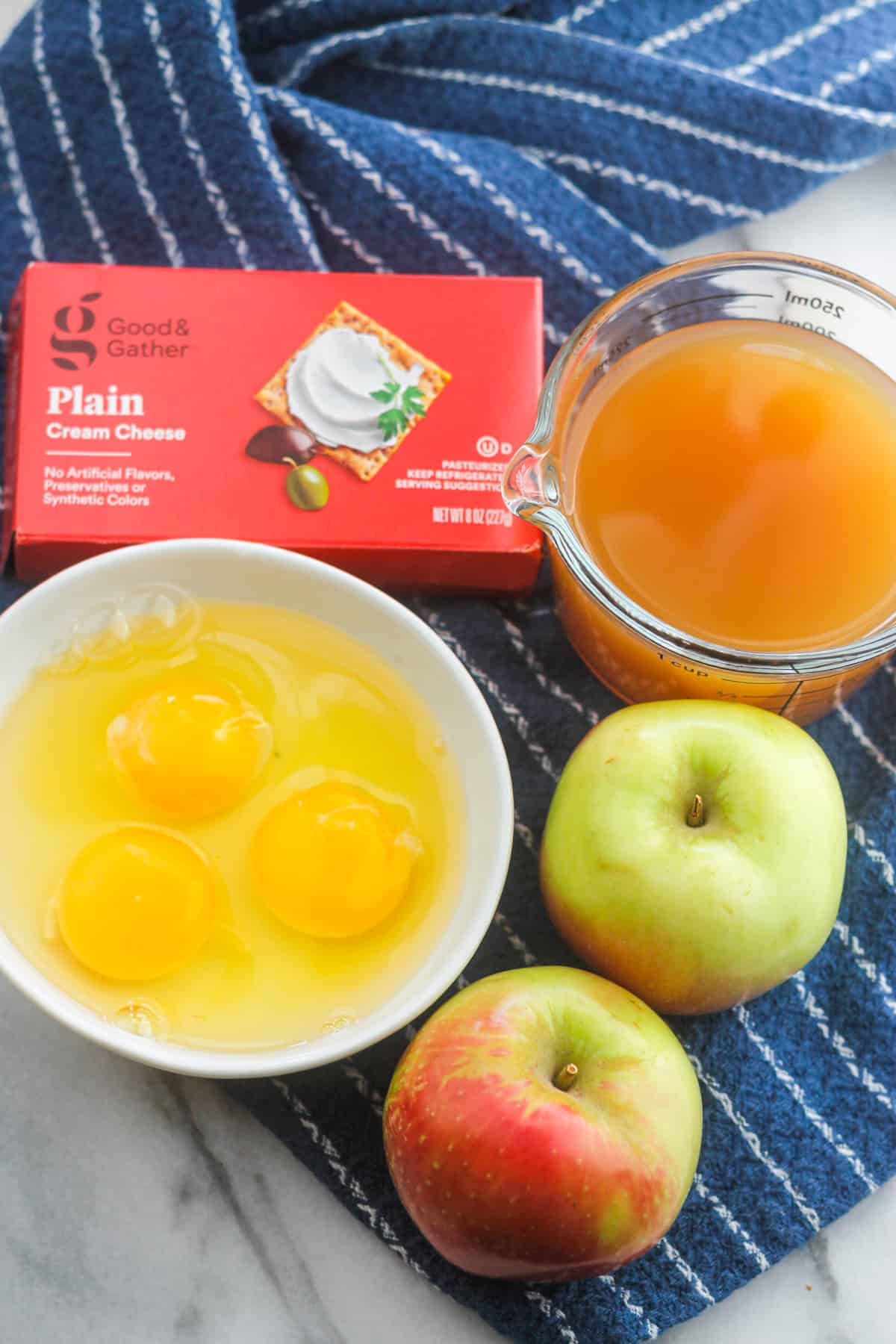 Clockwise from top left, boxed cream cheese, apple cider, two fresh apples, and bowl with three eggs.