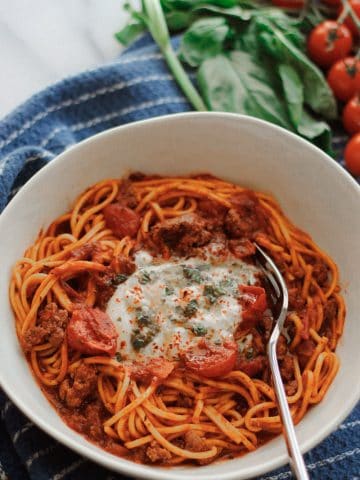 Overhead shot of Beefy pasta with tomatoes and burrata cheese garnished with lemon basil oil on blue towel with fresh basil and tomatoes above the bowl.