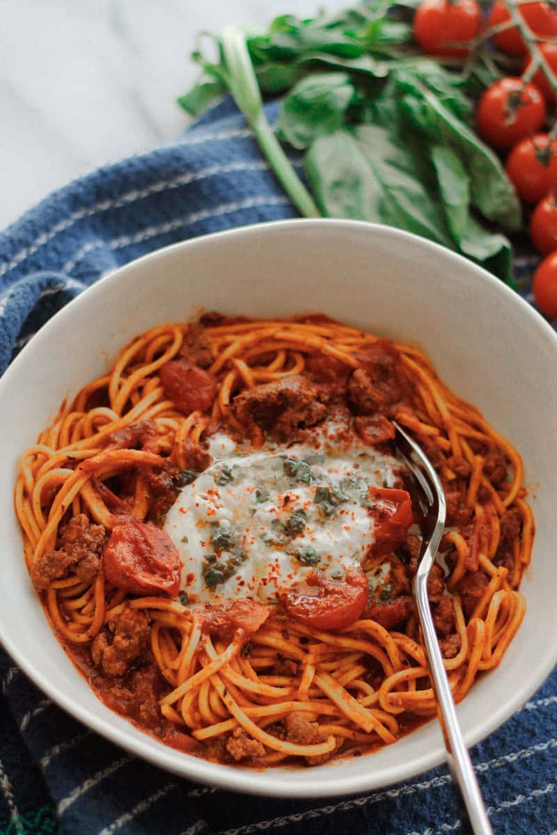 Overhead shot of Beefy pasta with tomatoes and burrata cheese garnished with lemon basil oil on blue towel with fresh basil and tomatoes above the bowl.