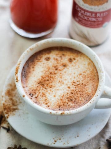 Overhead shot of Rooibos Chai Latte in a white cup and saucer sprinkled with cinnamon and a glass jar of Rooibos Chai tea concentrate and a tin of Rooibos tea behind the cup and saucer.
