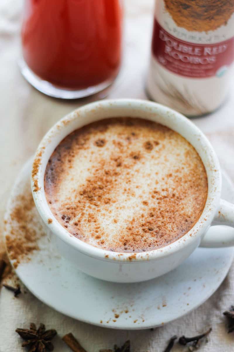 Overhead shot of Rooibos Chai Latte in a white cup and saucer sprinkled with cinnamon and a glass jar of Rooibos Chai tea concentrate and a tin of Rooibos tea behind the cup and saucer.