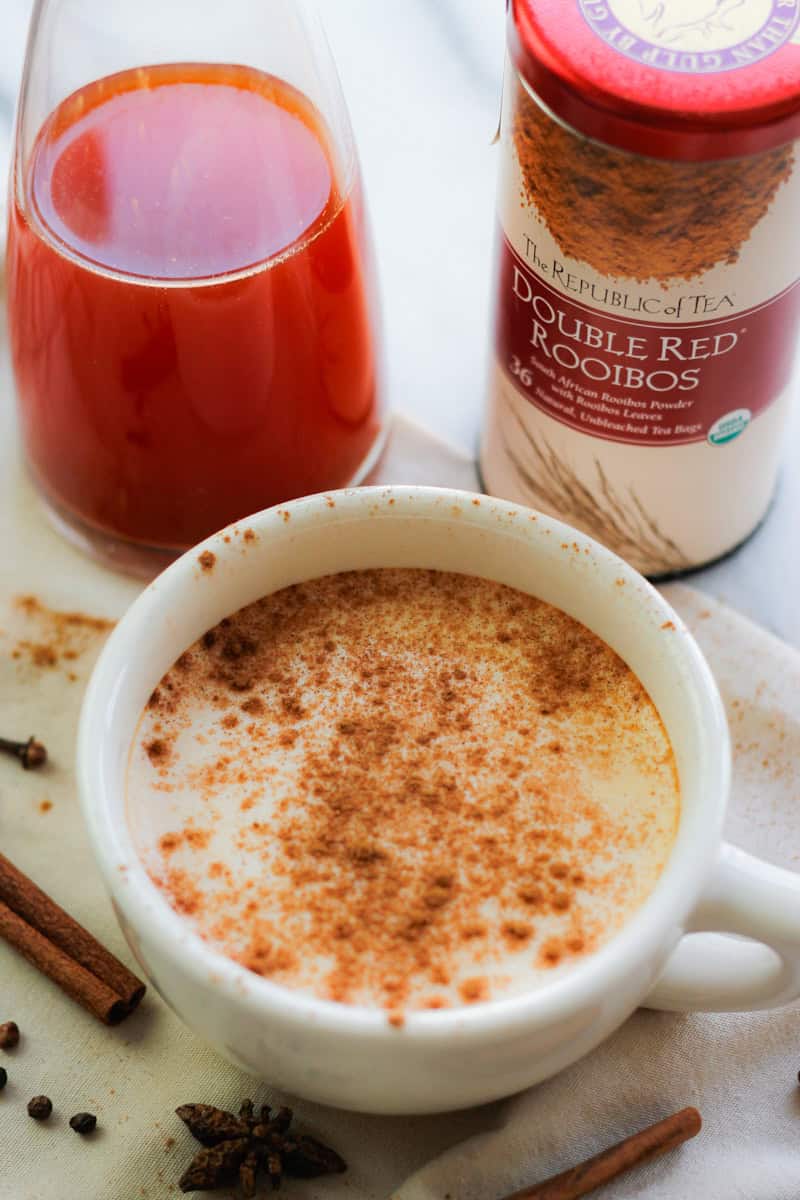 A cup of Rooibos Chai Latte with the Rooibos Chai tea concentrate and tin of Rooibos tea behind the cup of tea.