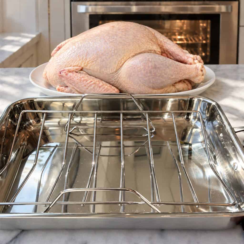 Roasting-
pan with rack on counter with uncooked turkey behind pan.